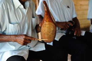 Instrument with rough surface being rubbed by a stick. Player of dark skin wearing traditional shirt.